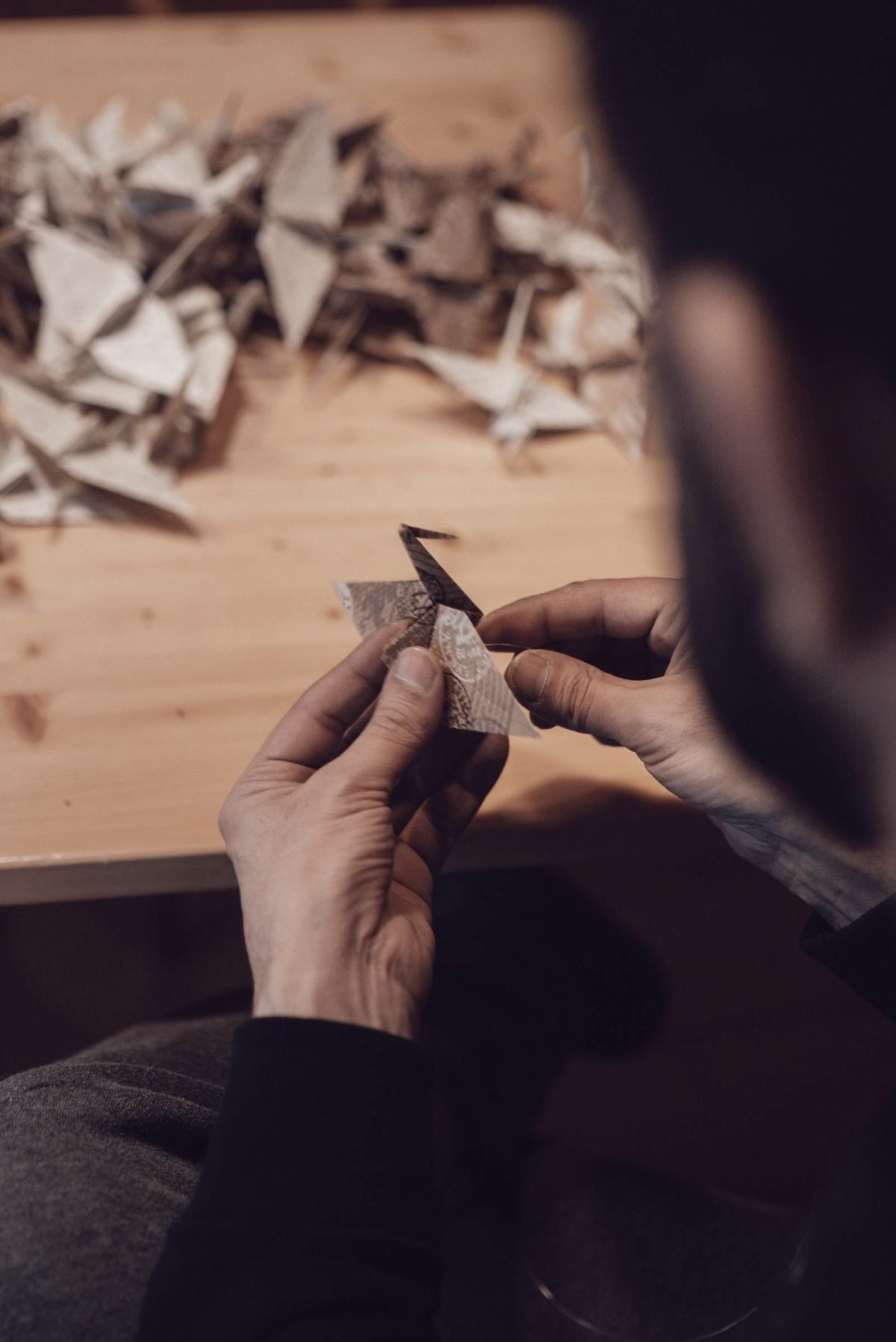 Hands pressing a fold into paper at a dark workbench, earlier folds scattered across the wood.